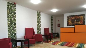 Un hall con sillas rojas, una mesa negra, un tapete a rayas de colores y una pared con estampado de hojas tropicales.