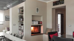 A modern kitchen with a built-in fireplace, a dining area with a black wicker table and chairs, and a bookshelf filled with books and decorative items. - Villa in the heart of Tuscany (Rigutino)