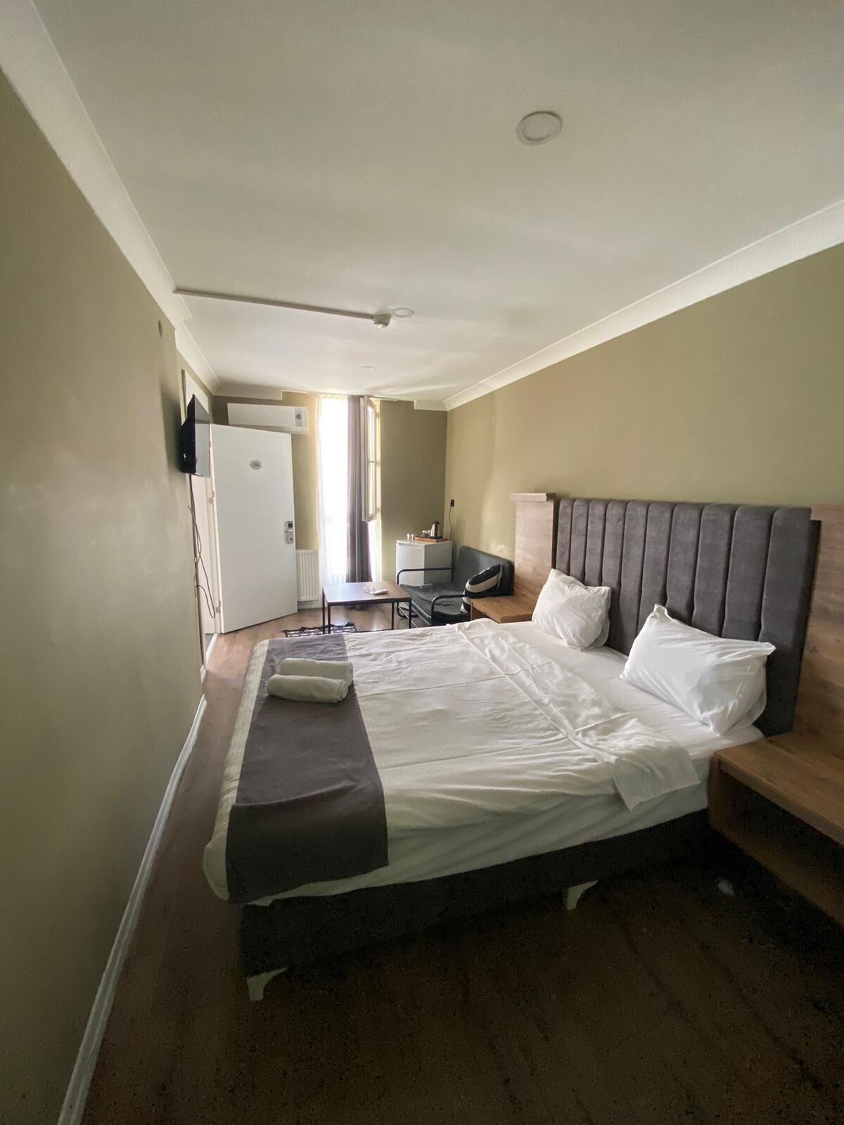 A hotel room with a large bed, a bedside table, a desk, and a chair. There is a window with curtains, a door, and an air conditioning unit on the wall.