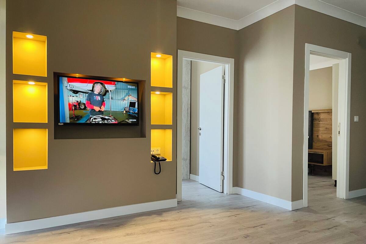 A modern living room with a flat-screen TV mounted on the wall, recessed lighting, and a wooden floor.