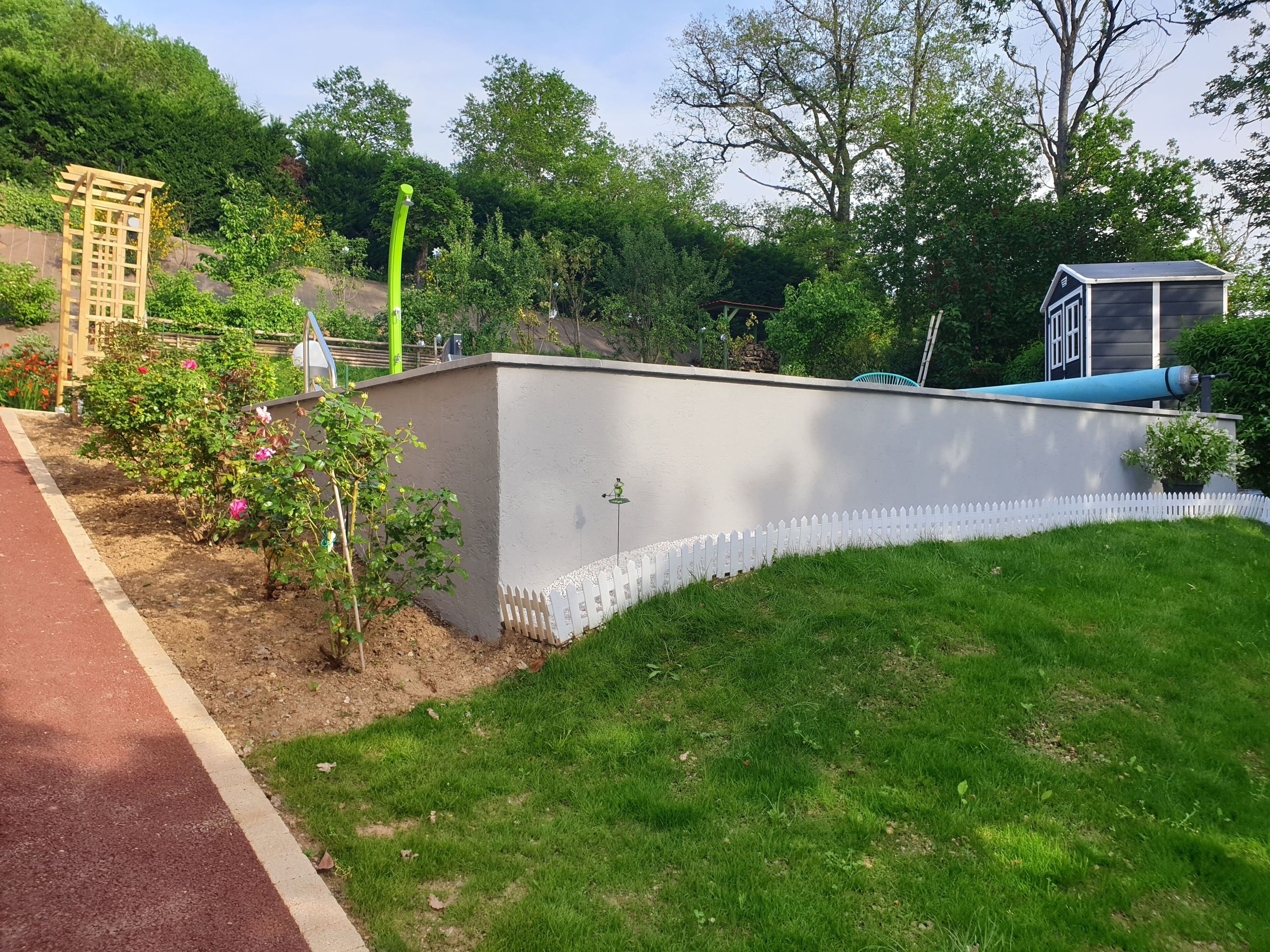 Un jardín con valla de piquetes blancos, una pérgola de madera y un cobertizo pequeño.