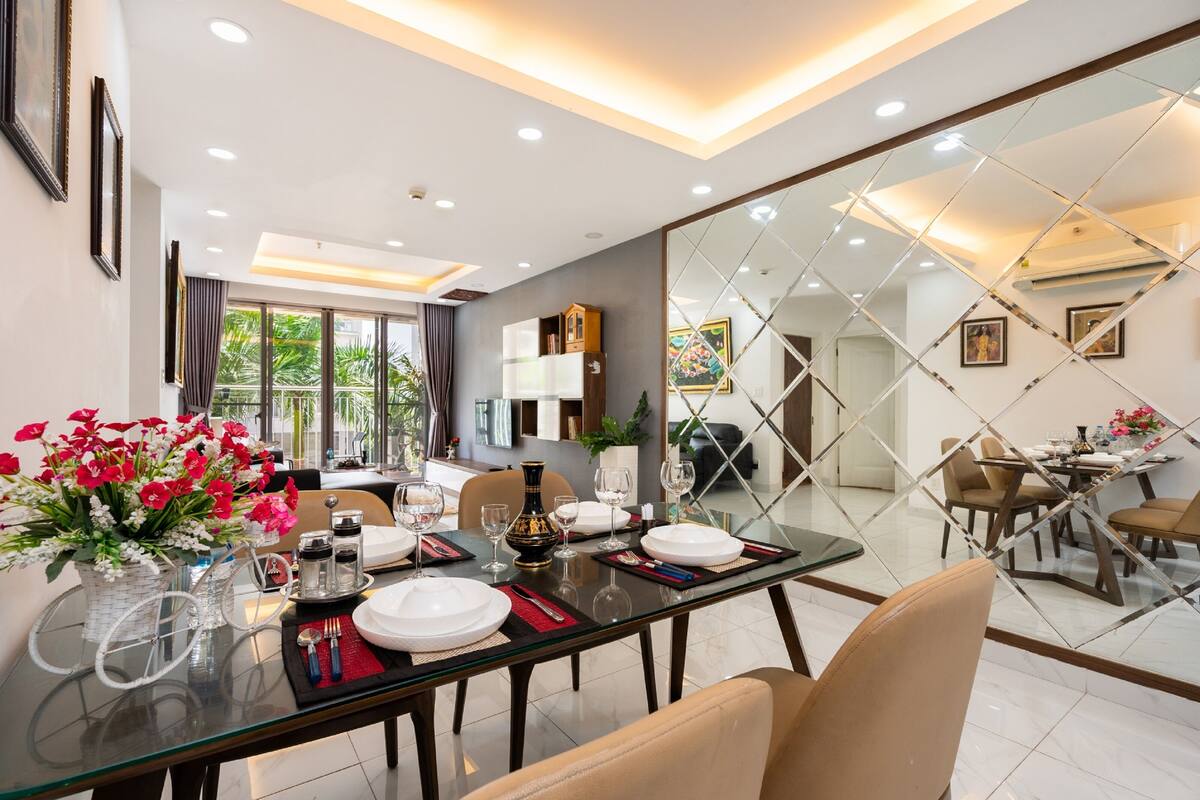 Un comedor con una mesa de vidrio dispuesta para cuatro personas, rodeada de sillas. Hay un jarrón con flores sobre la mesa, y la habitación tiene un diseño moderno con un espejo grande y vistas al exterior.