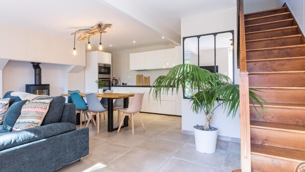A modern living room with a sofa, dining table, and chairs. There is a staircase and a kitchen in the background.