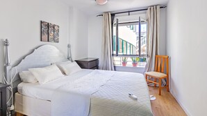 Un letto rifatto con lenzuola bianche, un comodino con una lampada, una sedia e una finestra con tende.