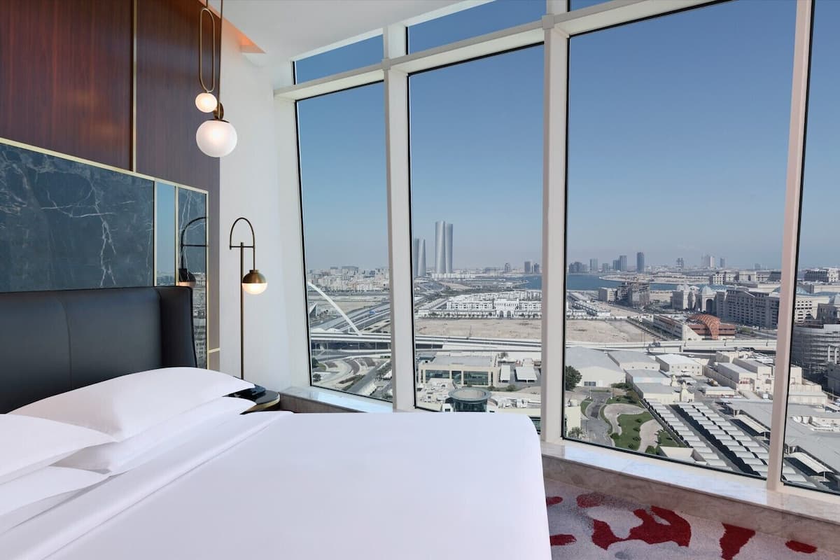 A modern hotel room with a large bed, a marble accent wall, and a city view through floor-to-ceiling windows.