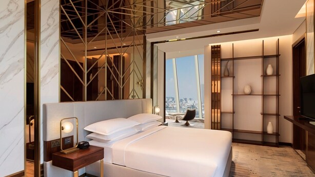 A modern hotel room with a large bed, a bedside table with a lamp, a TV, and a city view through large windows. - Velero Hotel Doha Lusail (Doha)