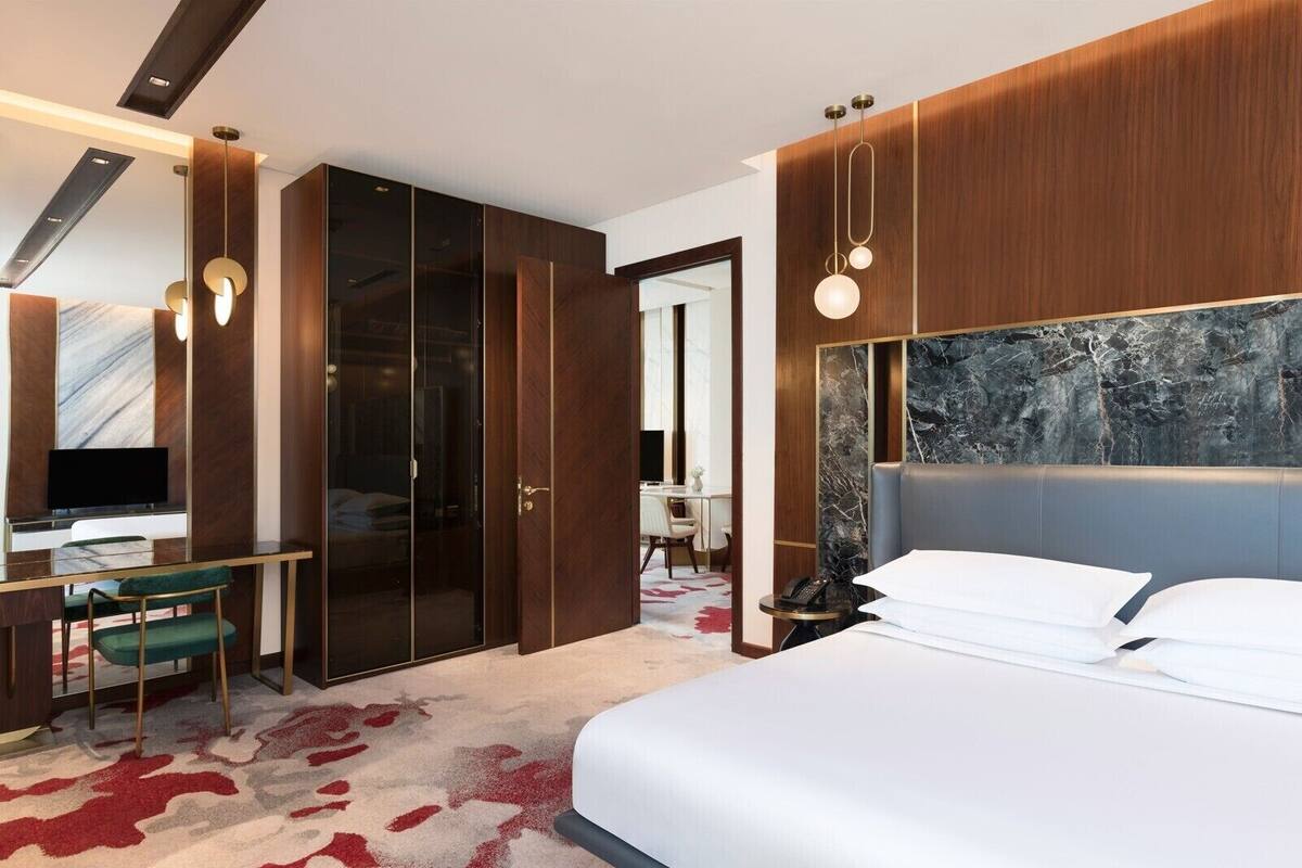 A modern hotel room with a large bed, a desk with a chair, a wardrobe, and a bathroom visible through a glass door.