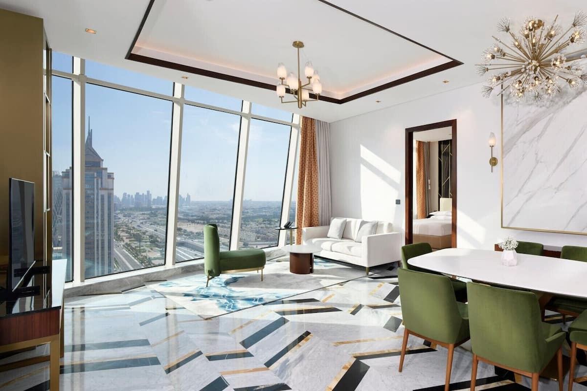 A modern hotel room with a large city view, a dining area with green chairs, and a marble floor.
