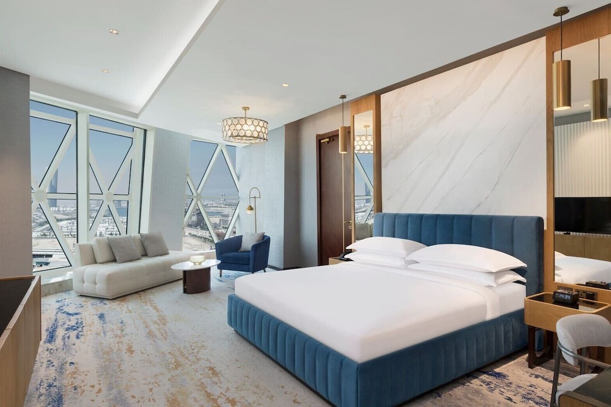 A modern hotel room with a large bed, a sofa, a TV, and a city view through large windows.
