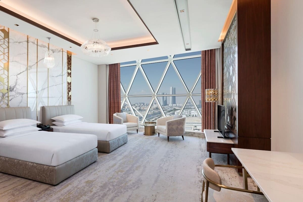 A modern hotel room with two beds, a desk, a TV, and a large window with a city view.