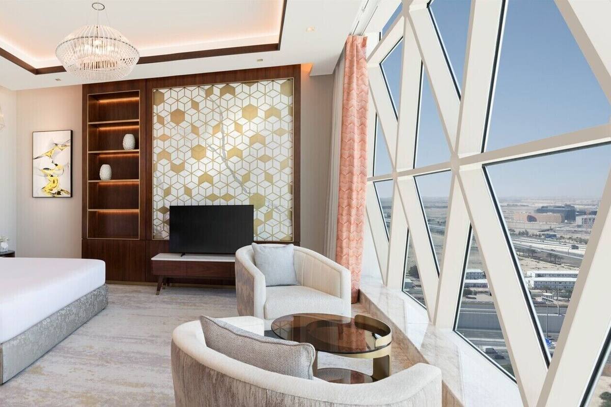 A modern hotel room with a large bed, a seating area, a TV, and a view of the city.