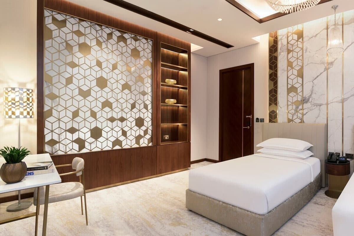 A modern hotel room with a large bed, a desk with a chair, a small table with a plant, and a decorative wall panel.
