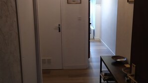 A hallway with a wooden floor, a door with a vent, a framed picture on the wall, and a table with a bowl and a plate. - Apartment (Crozon)