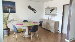 Een eetruimte met een ronde tafel, stoelen, een potplant en een stadsgezichtschilderij aan de muur.