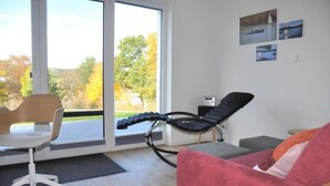 A modern room with a large sliding glass door leading to a balcony, a black massage chair, a red sofa, and a white office chair.