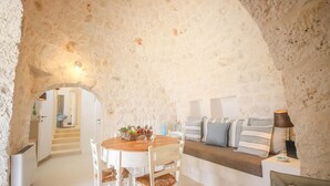 A cozy room with a stone archway, a round wooden table with chairs, a sofa with striped cushions, and a lamp on a side table. - TD Lamiamola Stylish Trulli Lamie With Pool (Ostuni)