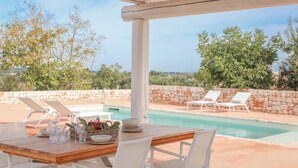 A poolside area with a wooden table set for dining, white lounge chairs, and a stone wall. - TD Lamiamola Stylish Trulli Lamie With Pool (Ostuni)