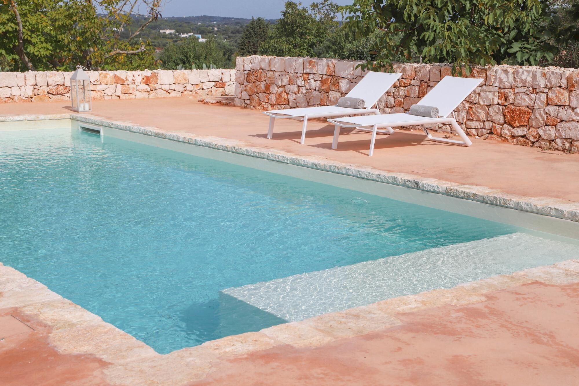 A rectangular pool with clear blue water, surrounded by a stone wall and two white lounge chairs.