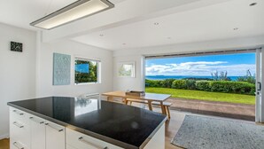 Una cocina moderna con una isla grande, un comedor con mesa y sillas, y un ventanal corredero que da a un jardín con vistas al mar.