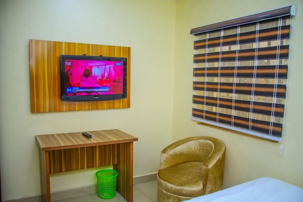 A small room with a television mounted on a wooden panel, a wooden desk, a green trash bin, a brown chair, and a window with striped blinds.