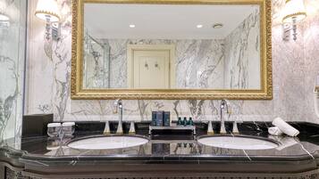 A bathroom with two sinks, marble walls, gold-framed mirror, and wall-mounted lights.