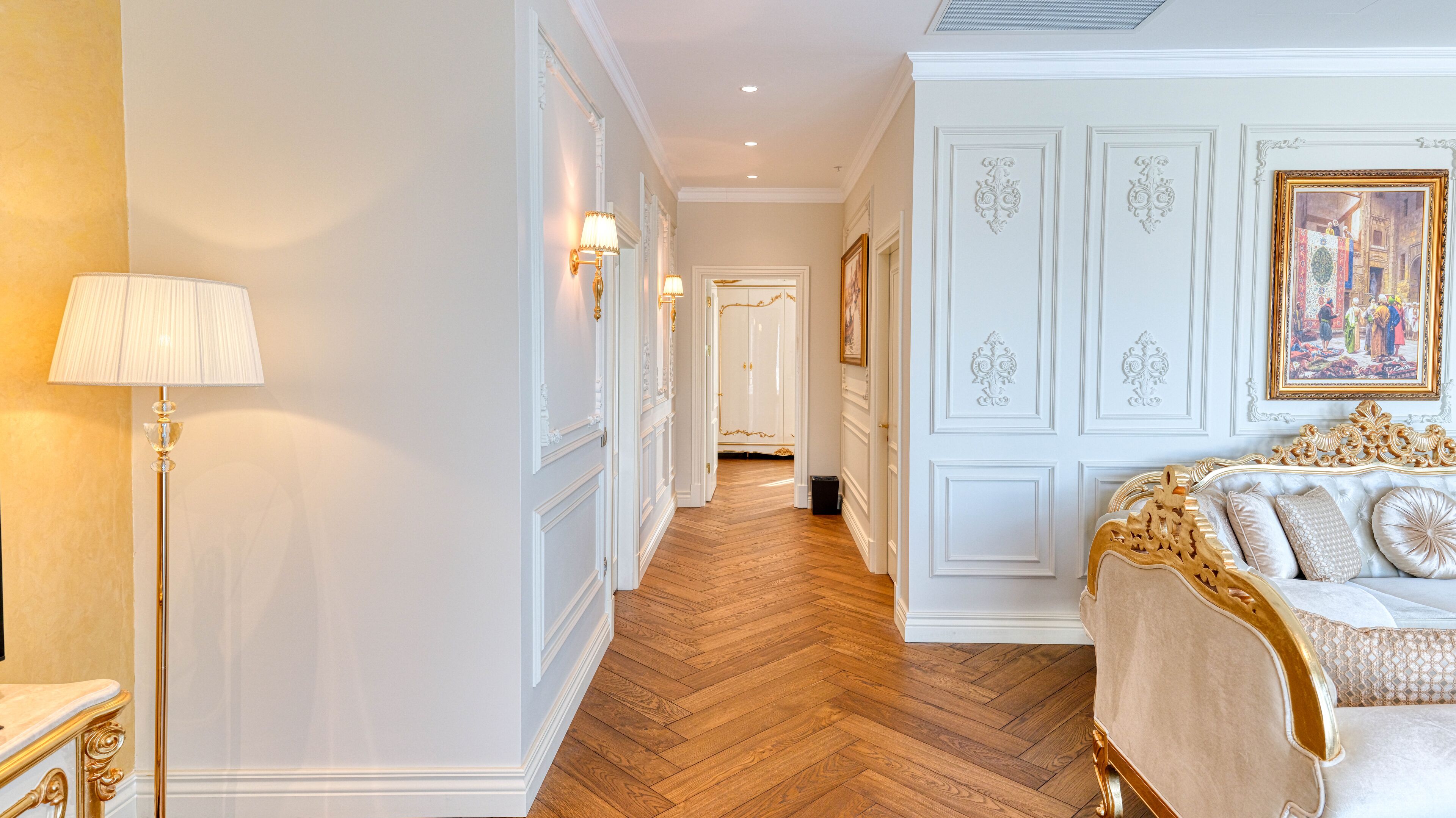 A hallway with a wooden floor, white walls, and a bed with gold detailing.