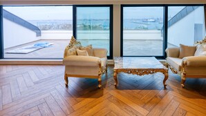 Um quarto espaçoso com duas sofás elaboradas e uma mesa de centro de mármore, grandes janelas do chão ao teto com vista para a cidade, e um piso de madeira em padrão herringbone.