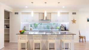 A modern kitchen with white cabinets, a central island with bar stools, a large window with a view of greenery, and pendant lighting. - Big Wave At Shoal Bay, 8 Achilles St - large home with ducted air con, Wi-Fi and Linen (Nelson Bay)
