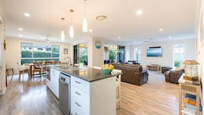 A spacious kitchen with a central island, a dining area with chairs, a living room with a sofa and a coffee table, and a view of greenery outside. - Big Wave At Shoal Bay, 8 Achilles St - large home with ducted air con, Wi-Fi and Linen (Nelson Bay)
