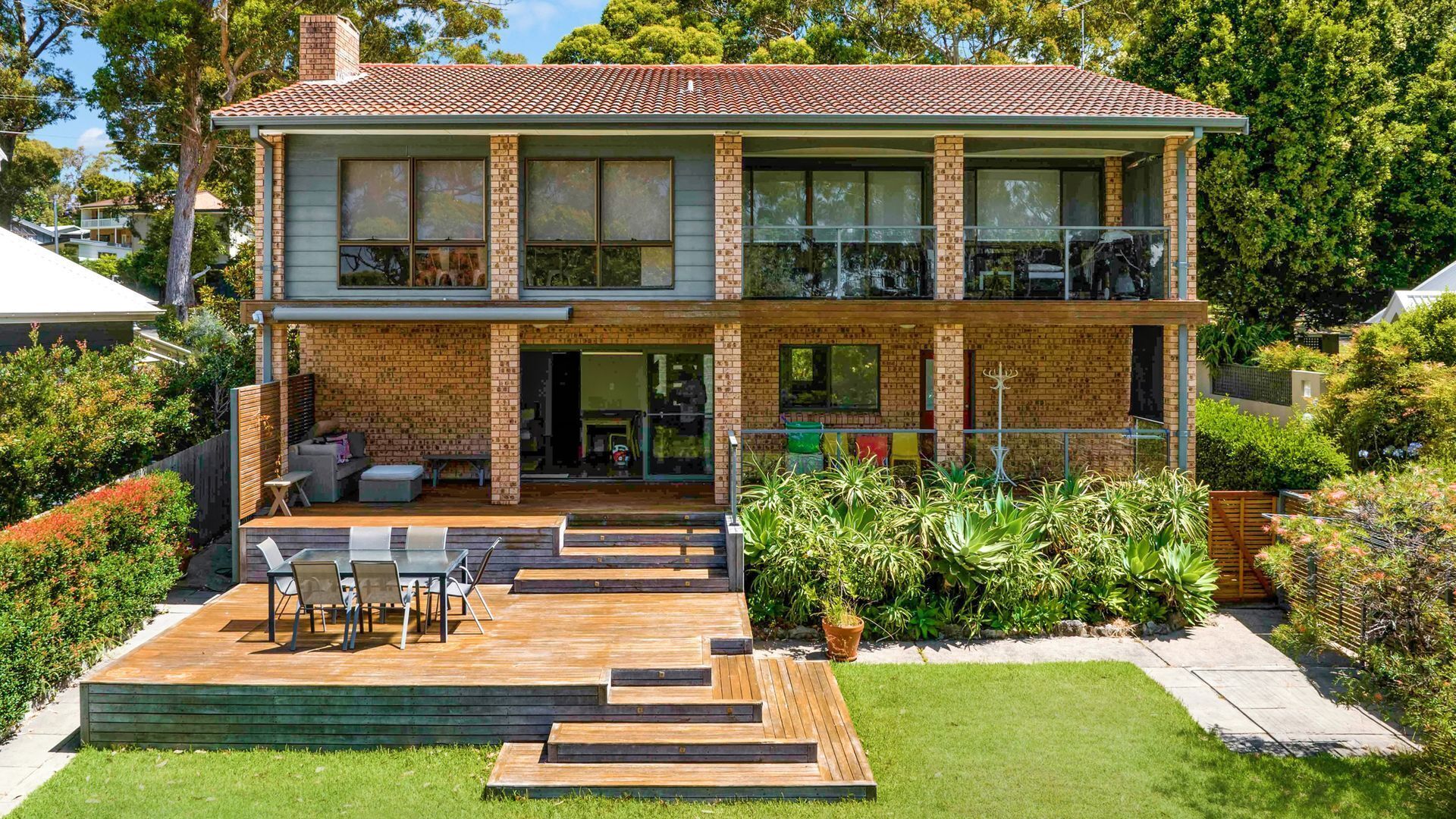 Une maison à deux étages avec une façade en briques, un balcon à l’étage supérieur et une terrasse en bois aménagée avec du mobilier d’extérieur.