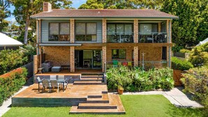 Een tweelaags huis met een bakstenen gevel, een balkon op de bovenverdieping en een houten terras met tuinmeubilair.