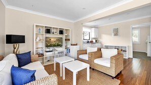Een woonkamer met een bank, twee witte tafels, rieten stoelen, een televisie en een keuken op de achtergrond.