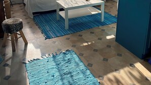 Un soggiorno con divano, tavolino da caffè e cuscini decorativi. Si nota una finestra con tende, un tappeto blu e un piccolo sgabello.