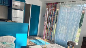 Una stanza con un letto, un tavolo bianco, una sedia in vimini, un divano blu e un angolo cottura con mobili azzurri, oltre a una finestra con tende.