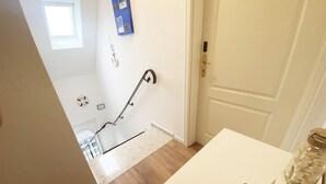 Una habitación con una puerta blanca, un ventanal pequeño, un objeto azul fijado a la pared y suelo de madera.