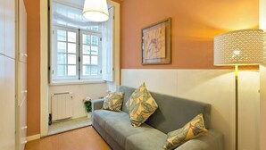 A cozy living room with a grey sofa, patterned cushions, a white radiator, and a window with white shutters. - PORTO CLÉRIGOS Apartments Daisy (1st) (Porto)