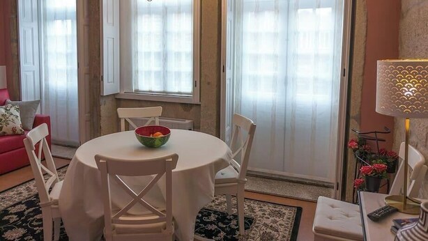 A dining area with a round table, white chairs, and a bowl of fruit. There is a red sofa and a lamp in the background.