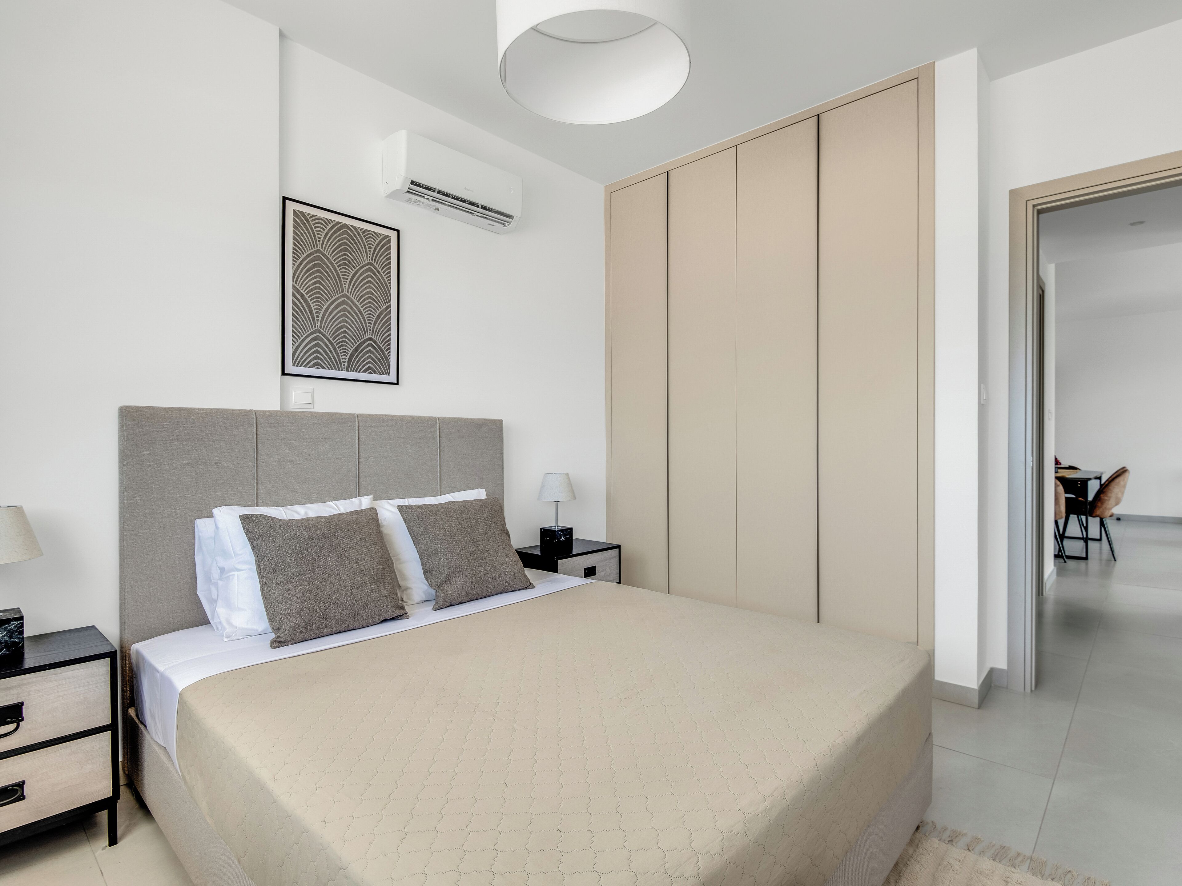 A modern bedroom with a large bed, a nightstand, a bedside table, a wall-mounted air conditioner, and a framed picture.
