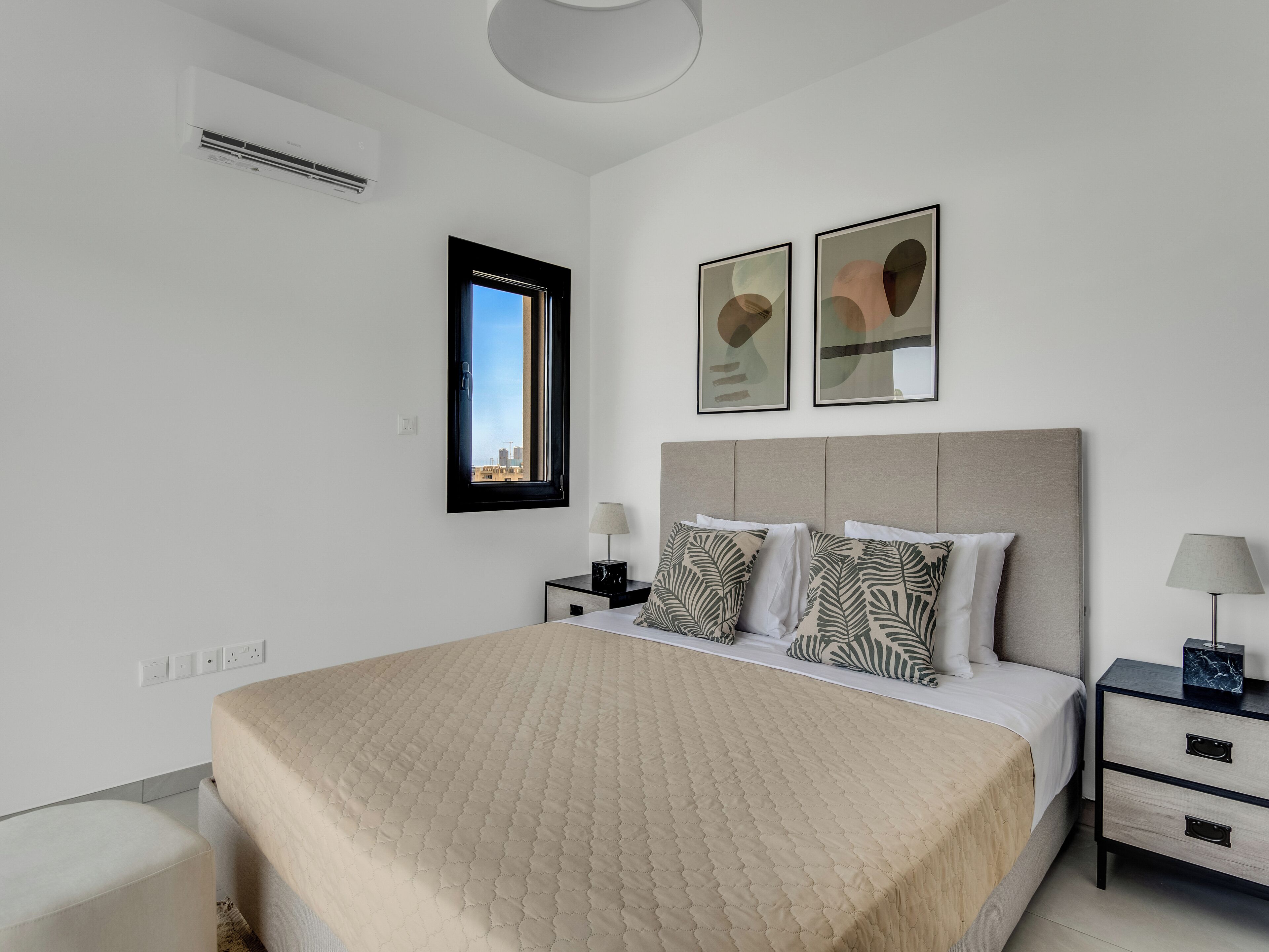 A modern bedroom with a large bed, a nightstand, a bedside table, a window, and two framed artworks on the wall.