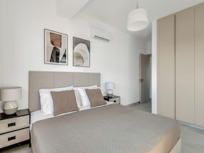 A modern bedroom with a large bed, bedside tables, a wardrobe, and two wall-mounted air conditioning units.