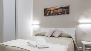 A neatly made bed with a beige bedspread and pillows, a white blanket folded at the foot, and a painting of a cityscape on the wall. - AdAstra b&b (Naples)