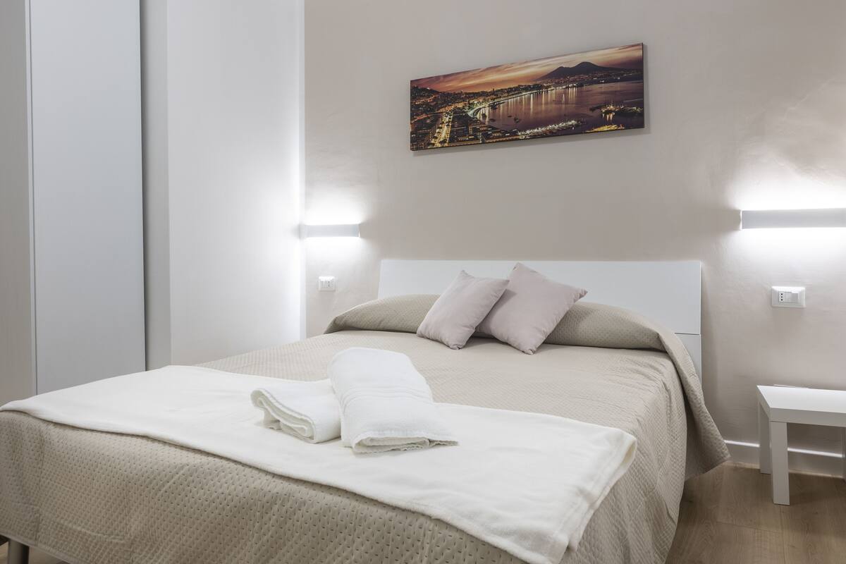 A neatly made bed with a beige bedspread and pillows, a white blanket folded at the foot, and a painting of a cityscape on the wall.