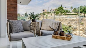 Un balcón con un sofá gris, una silla de mimbre y una mesa blanca con una bandeja de objetos.