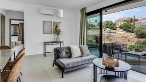 Una sala de estar moderna con un sofá gris, una mesa de centro redonda y un comedor con una mesa y sillas. Una ventana grande ofrece vistas del paisaje circundante.