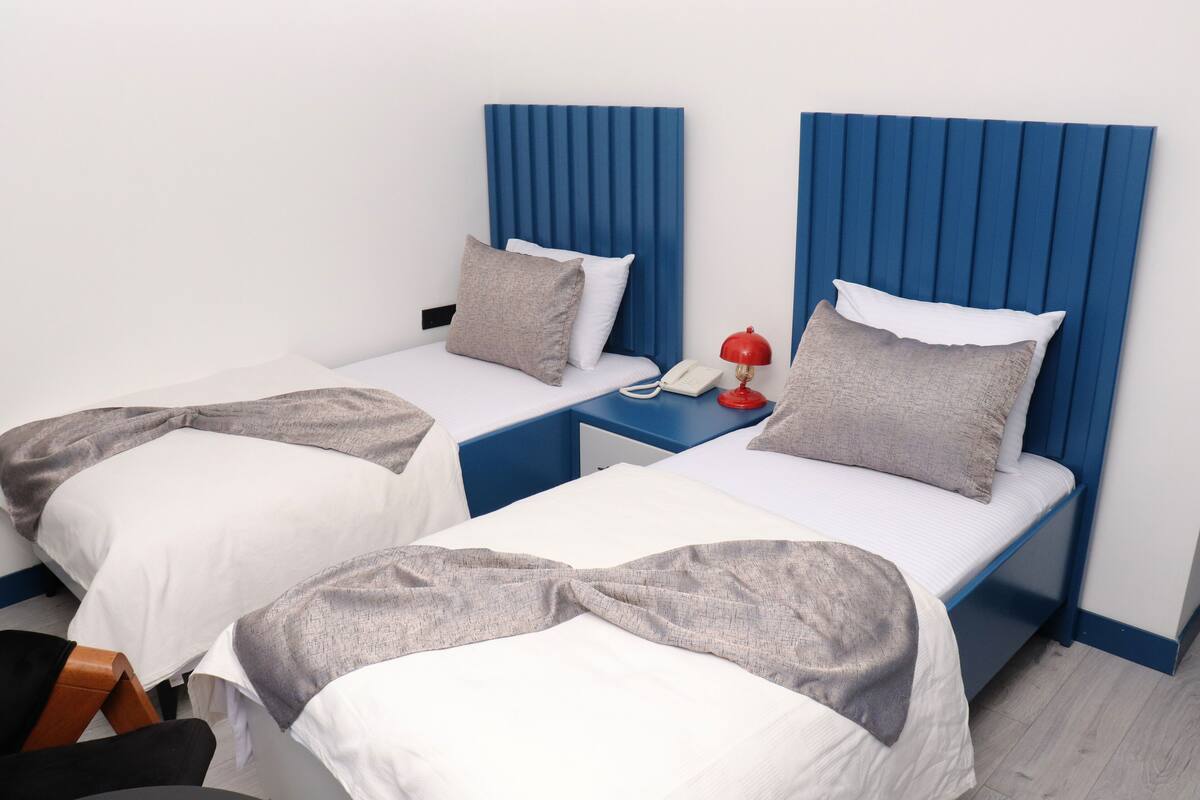 A hotel room with two single beds, each with a blue headboard, white bedding, and a grey throw blanket. There is a bedside table with a red lamp and a white phone.