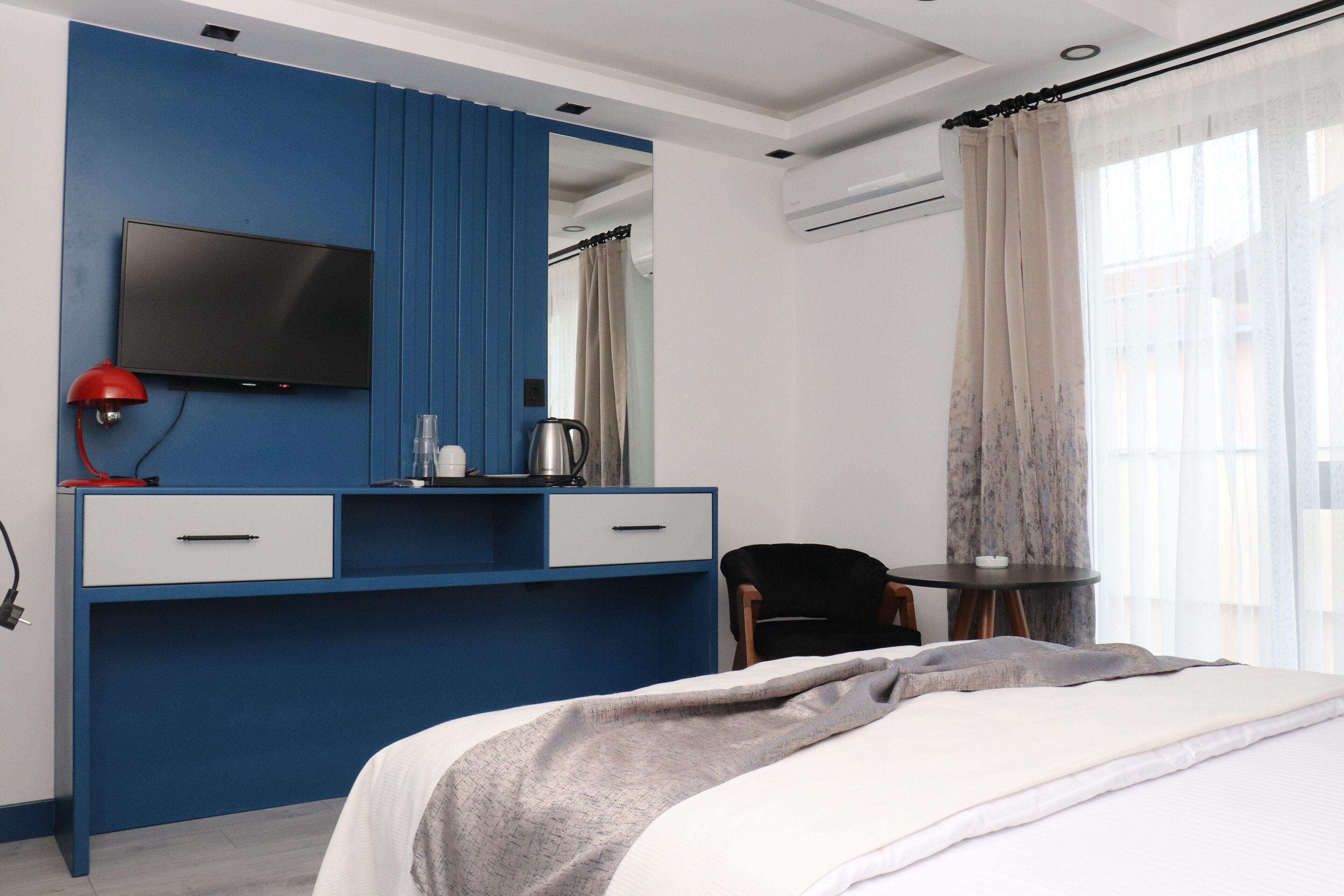A modern hotel room with a blue accent wall, a flat-screen TV, a desk with drawers, a chair, a bed with a grey blanket, and a window with sheer curtains.
