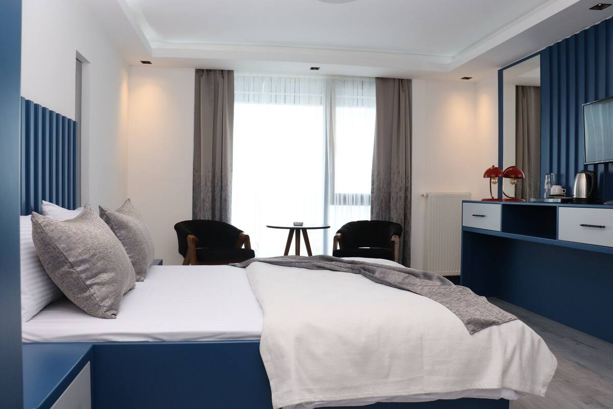 A modern hotel room with a large bed, two chairs, a small table, and a large mirror.