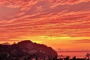 Un paisaje costero con un atardecer, una colina con edificios y una vista distante del mar.