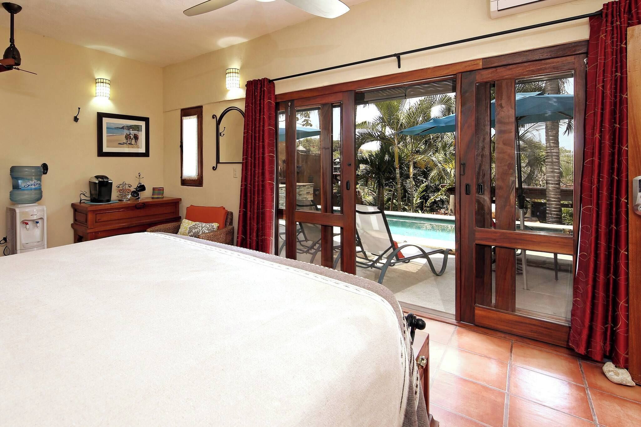 Un dormitorio con cama, cómoda de madera, dispensador de agua, un cuadro enmarco y una puerta corrediza de vidrio que da a un área exterior con piscina y sombrillas.