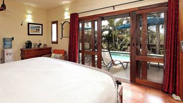 Un dormitorio con cama, cómoda de madera, dispensador de agua, un cuadro enmarco y una puerta corrediza de vidrio que da a un área exterior con piscina y sombrillas.